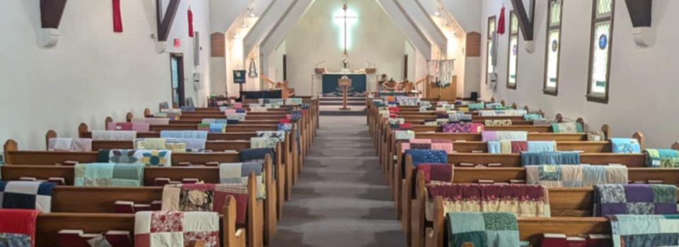 A picture of the sanctuary with many blankets laid over the pews to show our set up for our sewing group blanket sale to support CLWR